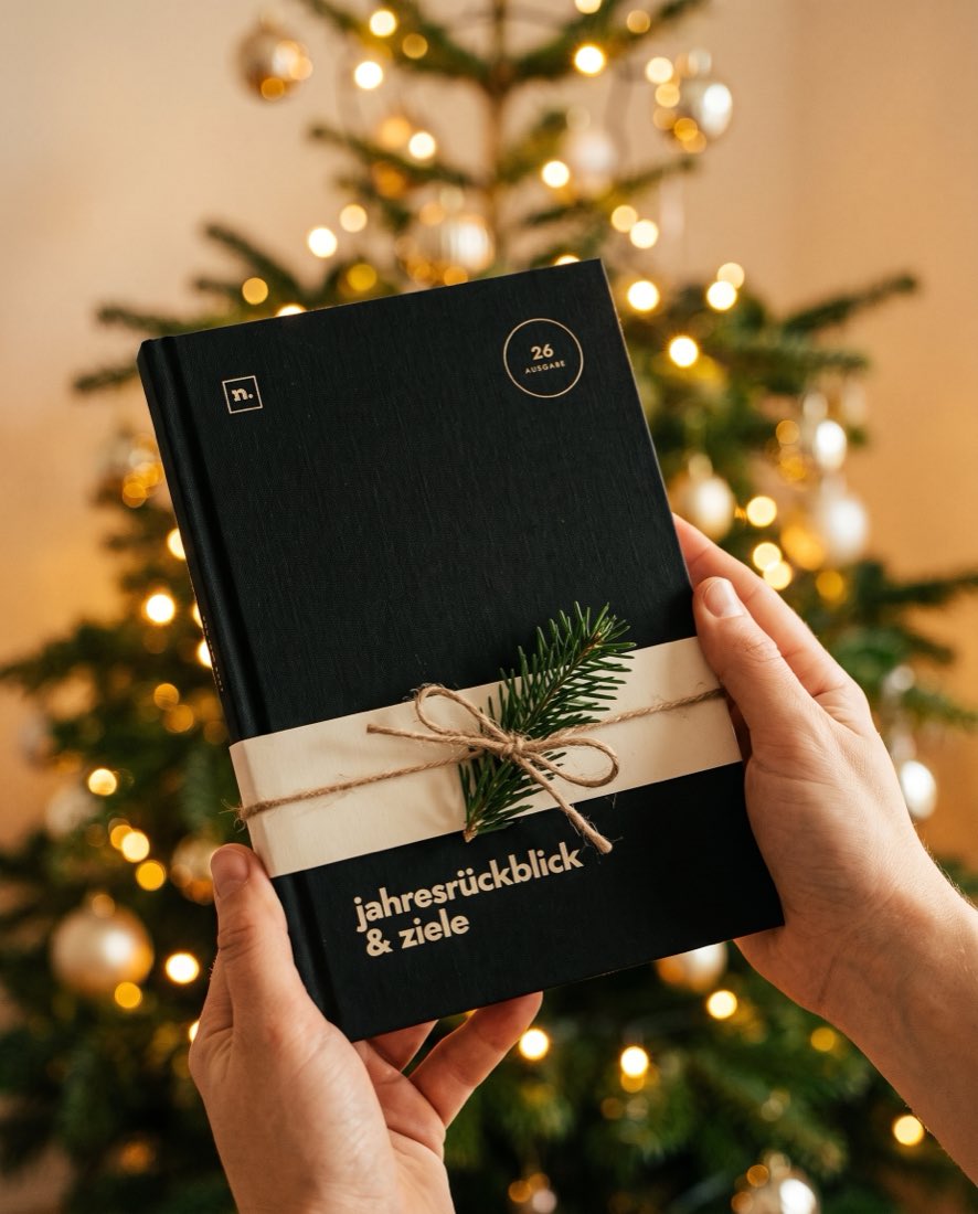 Zwei Hände halten die Ausgabe 26 mit Banderole und Tannenzweig vor einem geschmückten Weihnachtsbaum